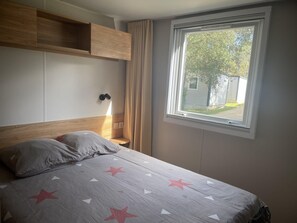 A bedroom with a bed, a nightstand, a lamp, and a window with a view of a building and trees. - Mobil-home 3 chambres 1 sdb (Mimizan)