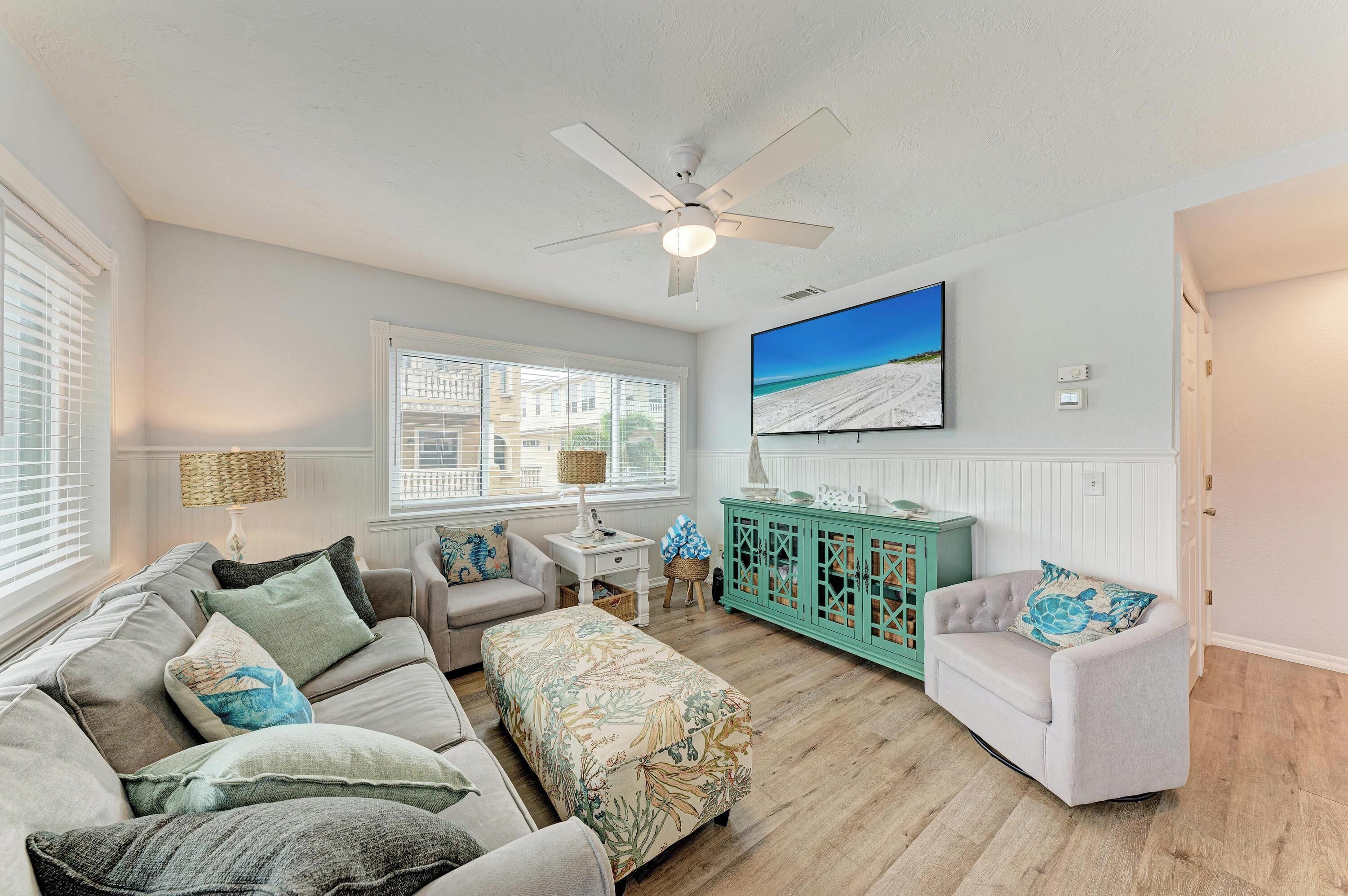 A living room with a ceiling fan, a large window with blinds, a television mounted on the wall, and a teal cabinet.