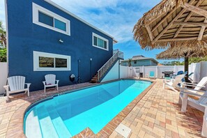 Piscina com borda azul, cercada por um pátio de tijolos, cadeiras brancas e uma estrutura de telhado de palha.