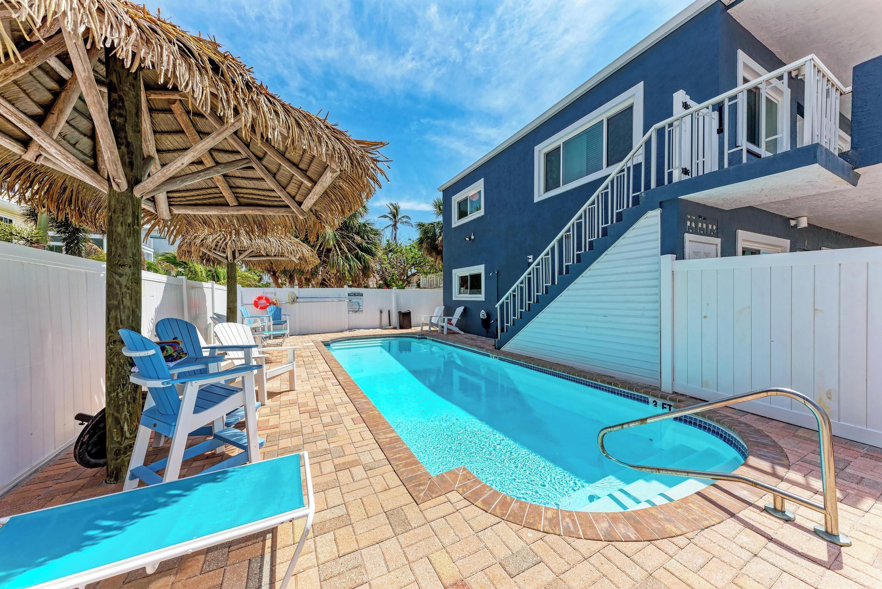 A swimming pool area with lounge chairs, a thatched umbrella, and a modern building with a blue facade.