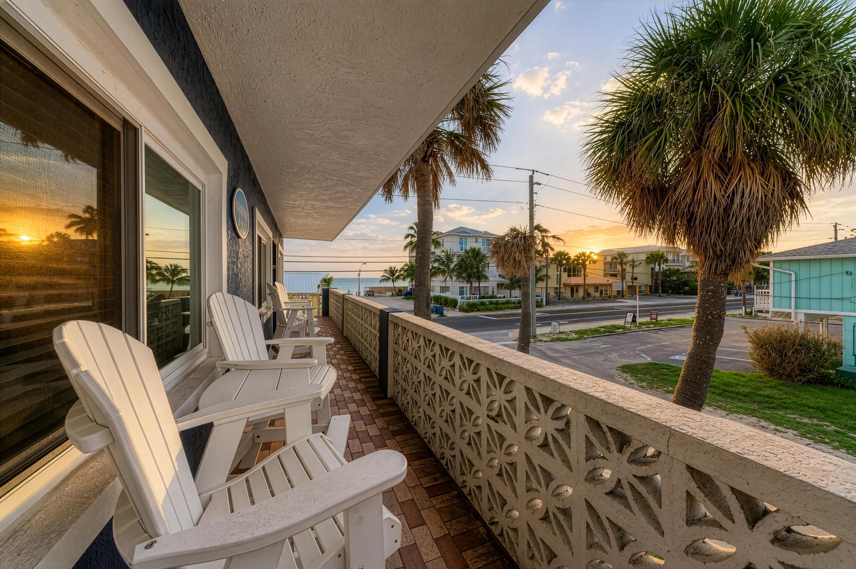 A balcony with two Adirondack chairs, a decorative fence, and a view of the ocean and palm trees.