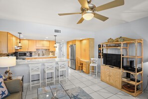 A modern kitchen with a central island, bar stools, a dining area, and a TV stand with a flat-screen TV and a sound system. - 🌅 Birdsong #26 — Just Steps to the Sand from this Treasure Island Delite (Treasure Island)