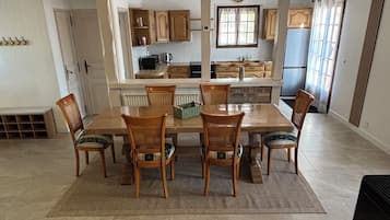 Een ruime eethoek met een lange houten tafel en stoelen, een keuken op de achtergrond en een open haard aan de rechterkant.