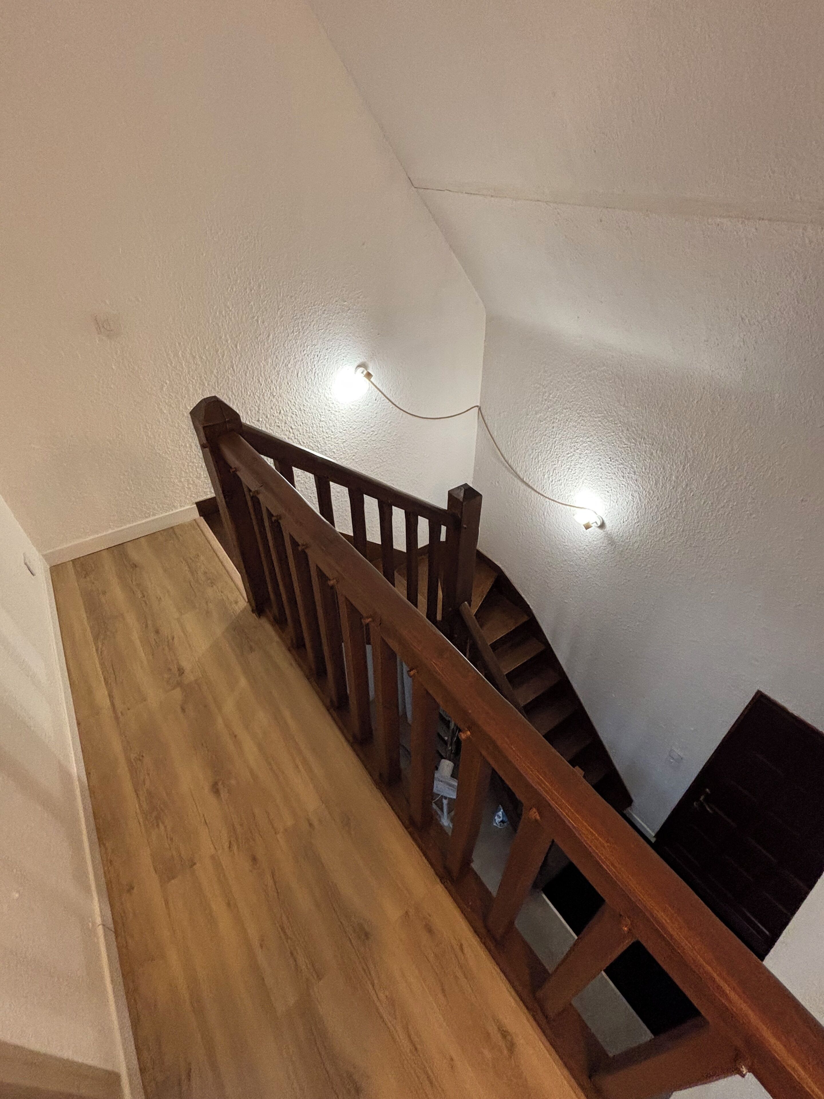 A wooden staircase with a handrail, white walls, and a light fixture on the wall.