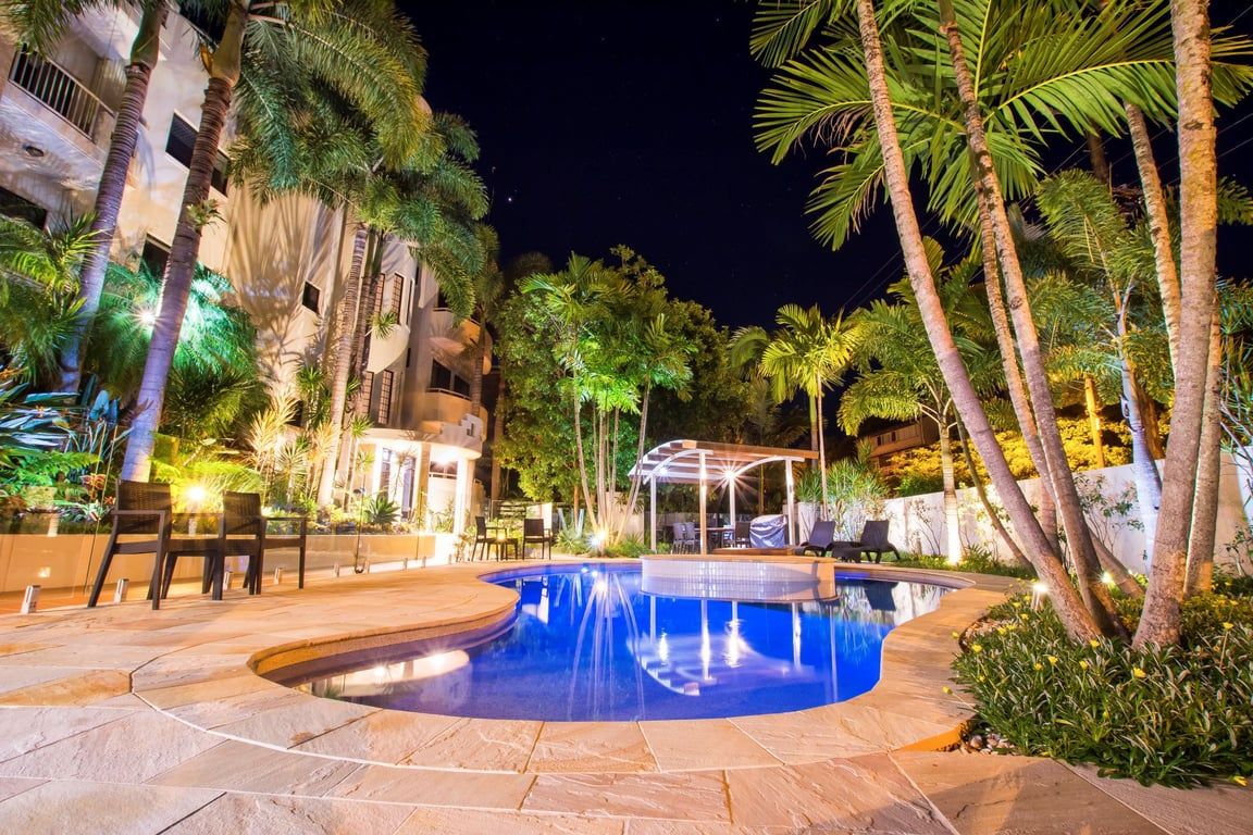 A nighttime swimming pool area with a curved design, surrounded by palm trees and outdoor seating.