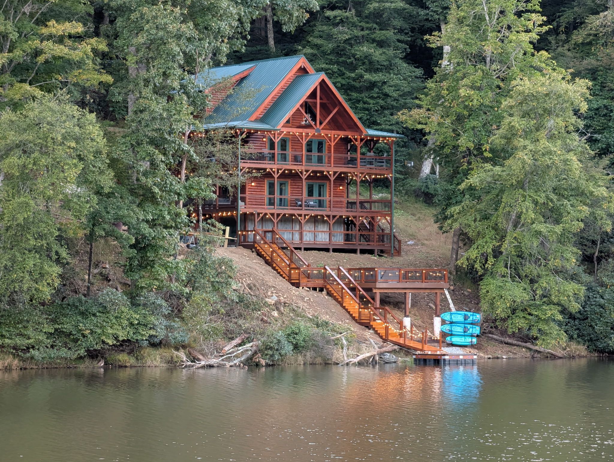 A multi-story wooden cabin with a metal roof, situated by a lake surrounded by dense forest.