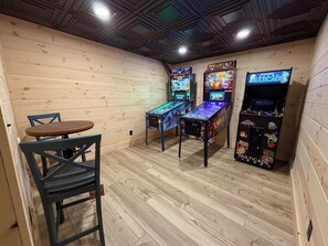 Una habitación con paredes de madera y techo inclinado, que presenta tres máquinas de arcade y una pequeña mesa con una silla.