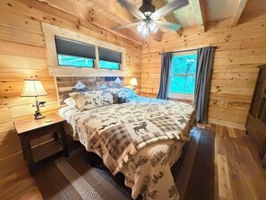 Un dormitorio de cabaña de madera con una cama, dos ventanas, un ventilador de techo y una mesita de noche con una lámpara.