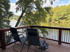 A wooden deck with two folding chairs overlooking a calm lake, surrounded by trees and a distant building. - Log Cabin on the Lake with Private Dock-Hot Tub-Fireplace-Arcade-Close to town! (West Jefferson)