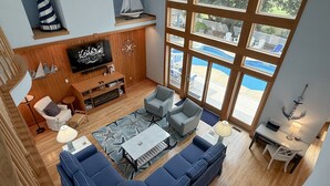 A spacious living room with a large window, a television, and a sailboat decoration. - Beach ParTee Time: Soundfront, Private Pool, Hot Tub, Community Amenities (Corolla)