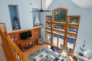 A spacious living room with a large window, wooden flooring, a television, and a sailboat decoration. - Beach ParTee Time: Soundfront, Private Pool, Hot Tub, Community Amenities (Corolla)