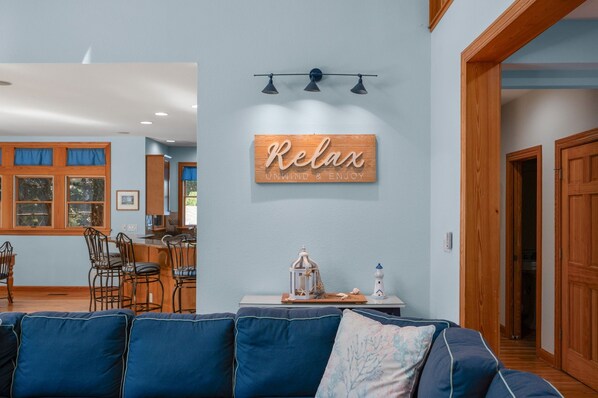 Un salon avec un canapé bleu, un panneau en bois sur lequel est écrit « Détendez-vous et profitez de l’instant présent », et un coin repas avec des chaises et une table.
