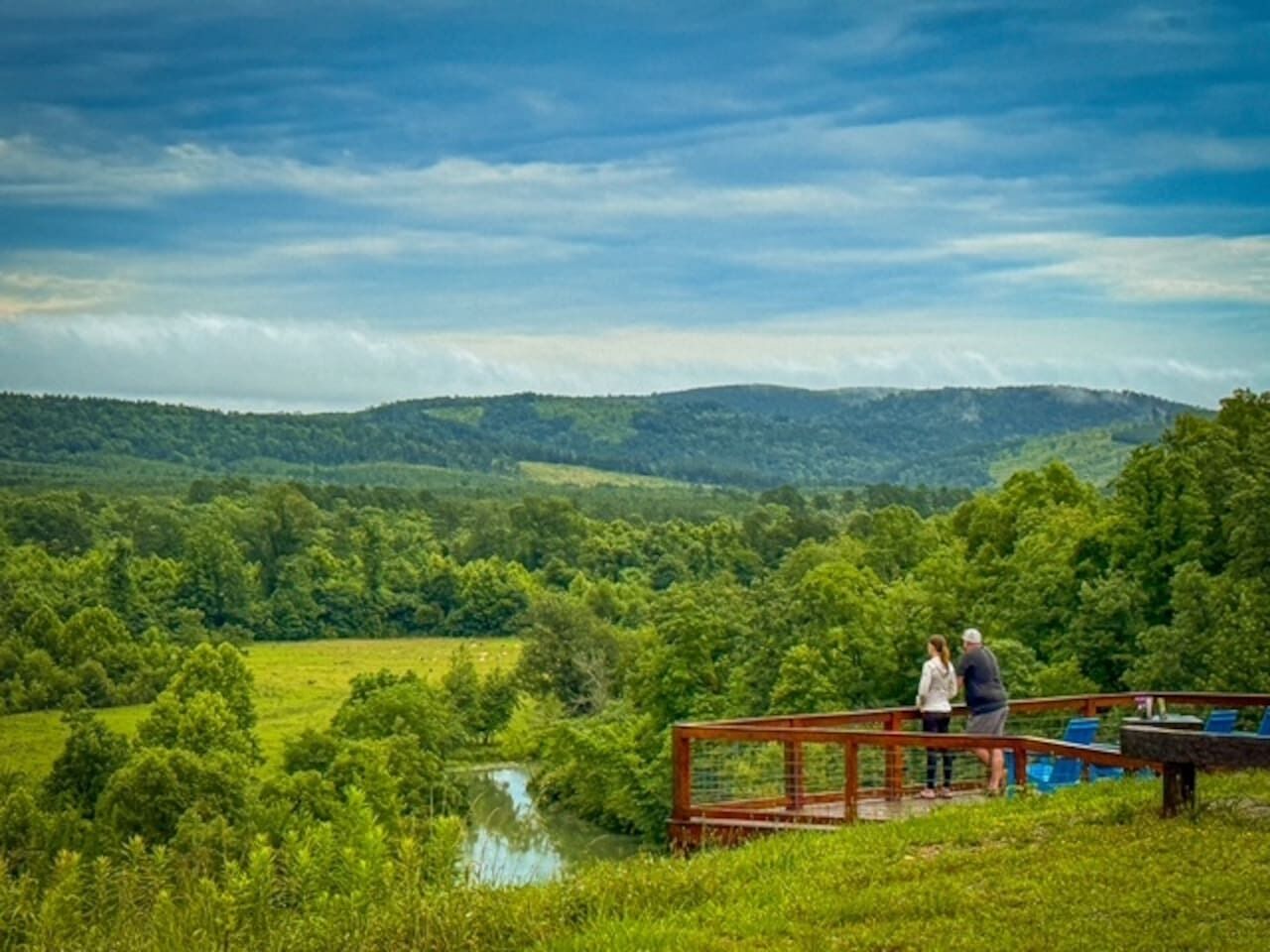 A couple standing on a wooden deck overlooking a lush green landscape with a river and hills in the distance.