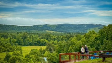 A couple standing on a wooden deck overlooking a lush green landscape with a river and hills in the distance.