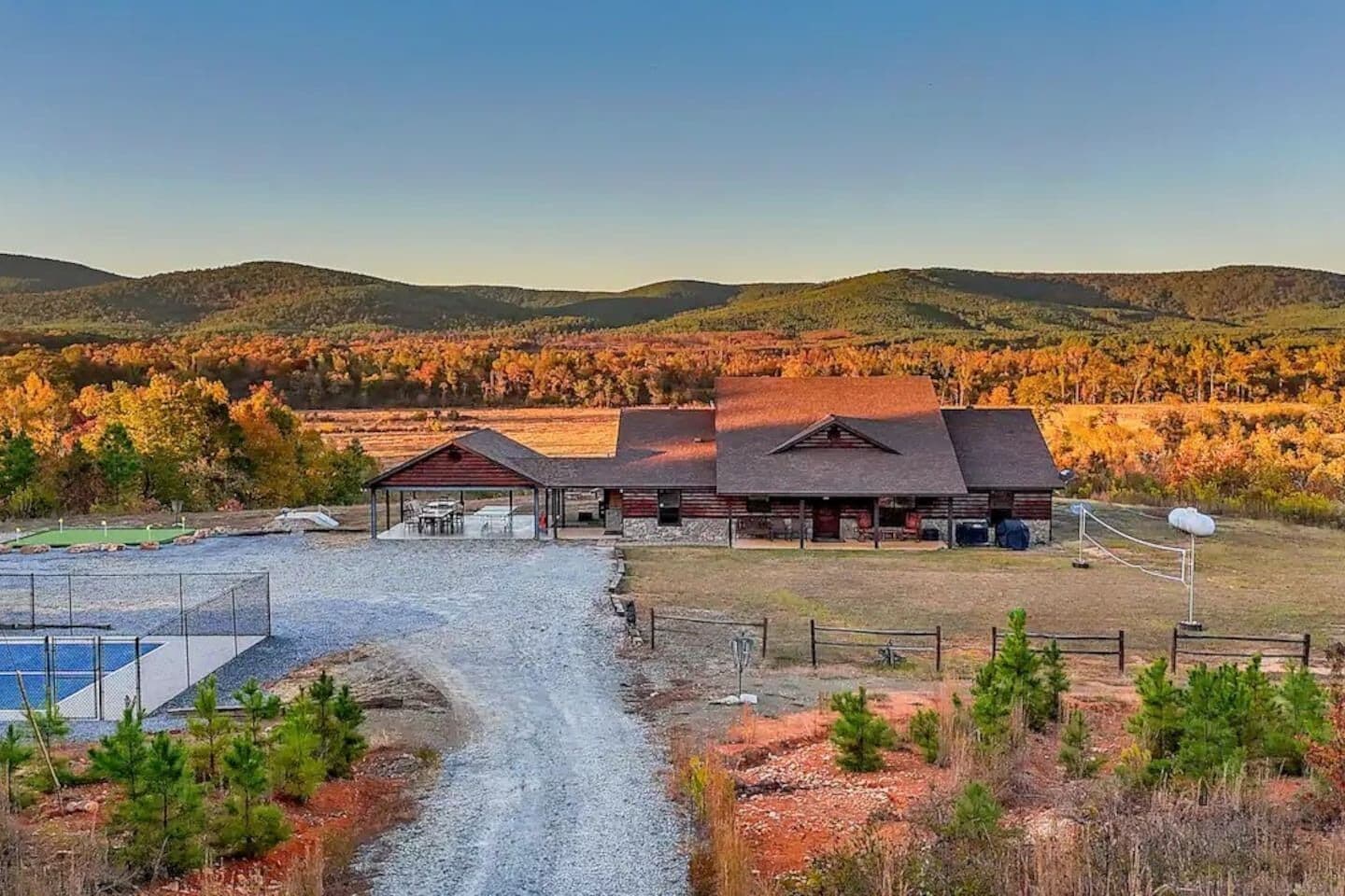A rural property with a large barn, a covered outdoor seating area, and a gravel driveway leading to a fenced sports court.