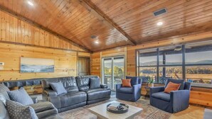 A spacious room with a wooden ceiling, leather sofas, a coffee table, and a large window offering a view of the outdoors. - River Cabin with Pickleball and Kayaks (Smithville)