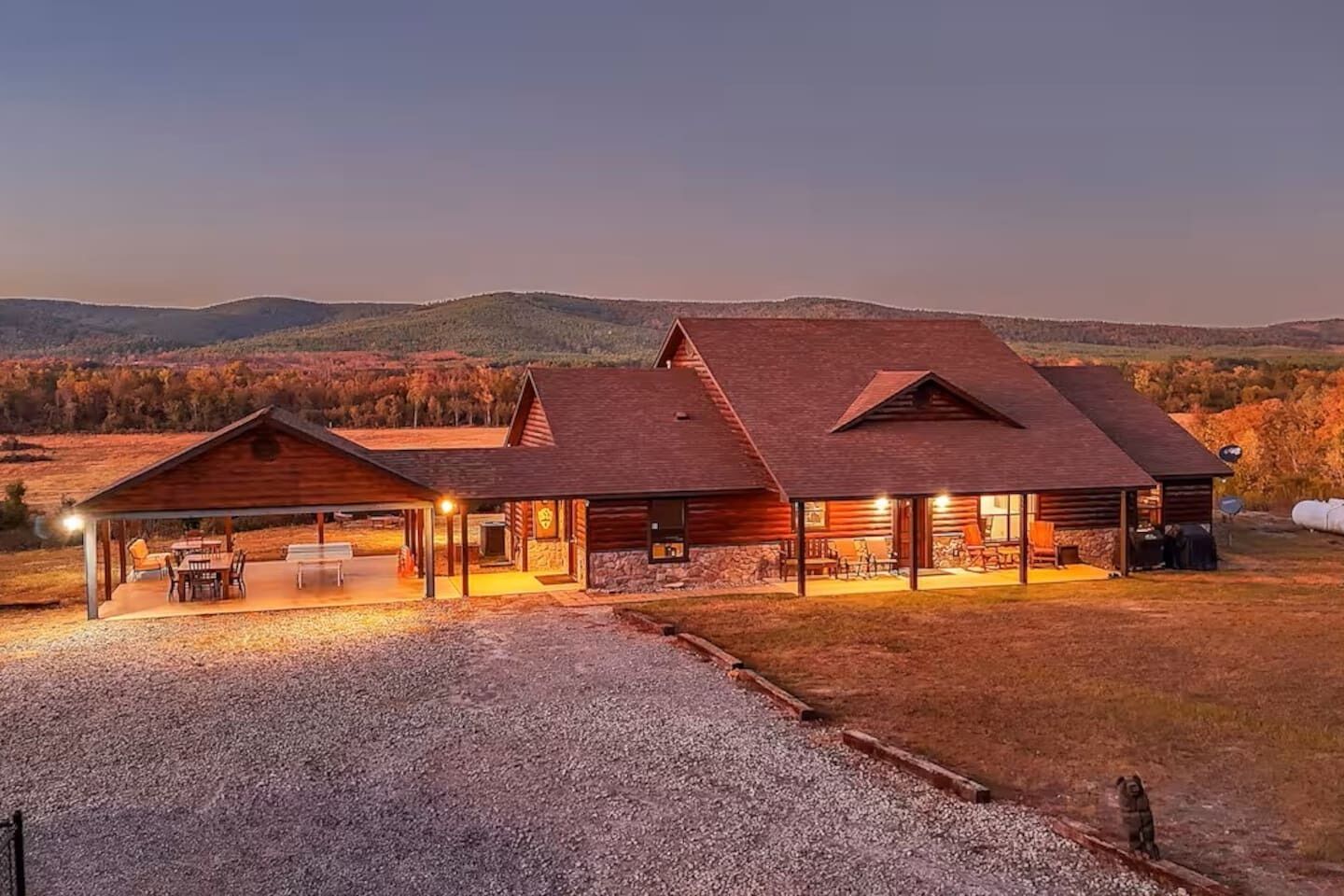 A log cabin with a covered patio and a stone foundation, set in a rural landscape with hills in the background.