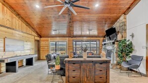 A spacious room with a wooden ceiling, a large window, a fireplace, and a seating area with a sofa and a dining table. - River Cabin with Pickleball and Kayaks (Smithville)