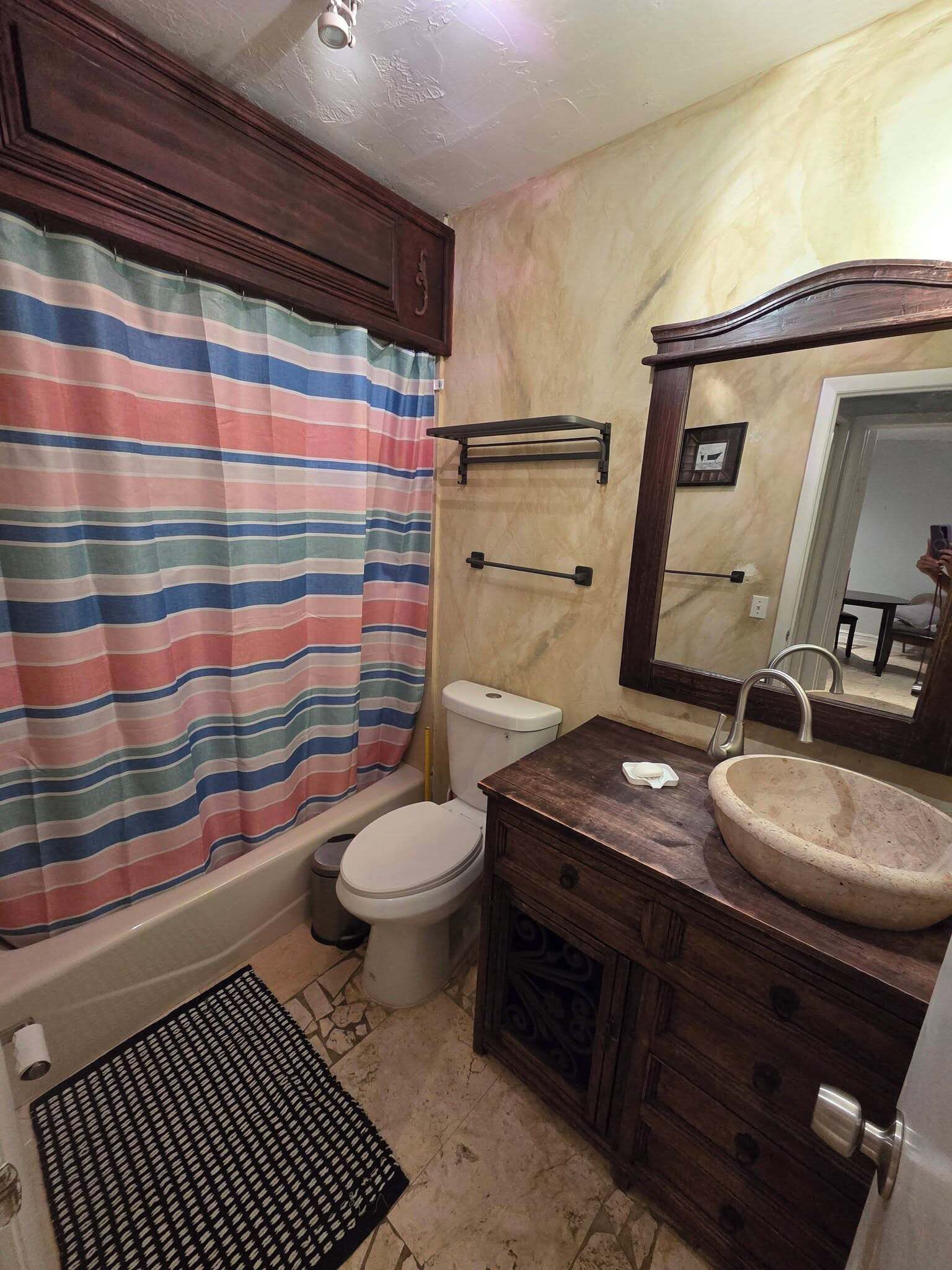 Een badkamer met een gestreept douchegordijn, een toilet, een wastafel met spiegel en een houten opbergmeubel.