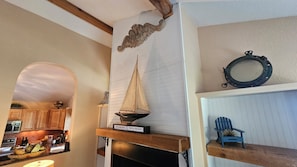 A room with a wooden mantelpiece, a small sailboat model, a blue Adirondack chair, and a kitchen visible through an archway.