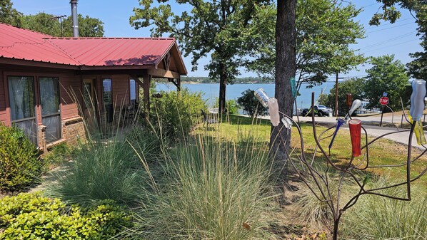 A wooden cabin with a red roof, surrounded by grass and trees, overlooking a body of water.