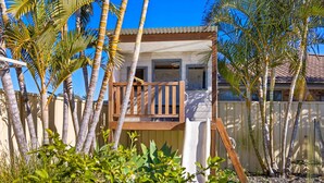Uma casa com deck de madeira, cercada por palmeiras e um quintal com cerca.