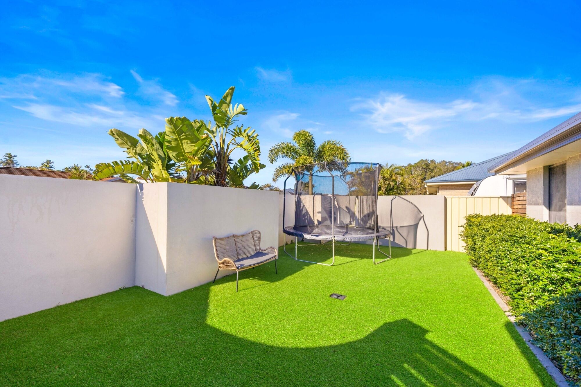 A backyard with artificial grass, a trampoline, a wicker chair, and a white wall.