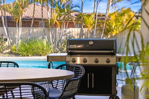 An outdoor grill with a gas tank, a table, and chairs, near a swimming pool.
