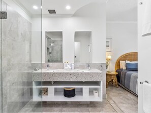 A modern bathroom with a glass-enclosed shower, a double sink vanity, and a bed with a headboard in the adjacent room. - Deluxe Three-Bedroom Apartment Apt 7 (Mooloolaba, Sunshine Coast)