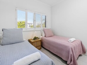 A bedroom with two beds, a window with blinds, a bedside table with a lamp, and a small plant. - Deluxe Three-Bedroom Apartment Apt 7 (Mooloolaba, Sunshine Coast)