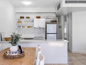 A modern kitchen with white cabinetry, a built-in refrigerator, and a dining area with a wooden table and chairs. - Deluxe Three-Bedroom Apartment Apt 7 (Mooloolaba, Sunshine Coast)