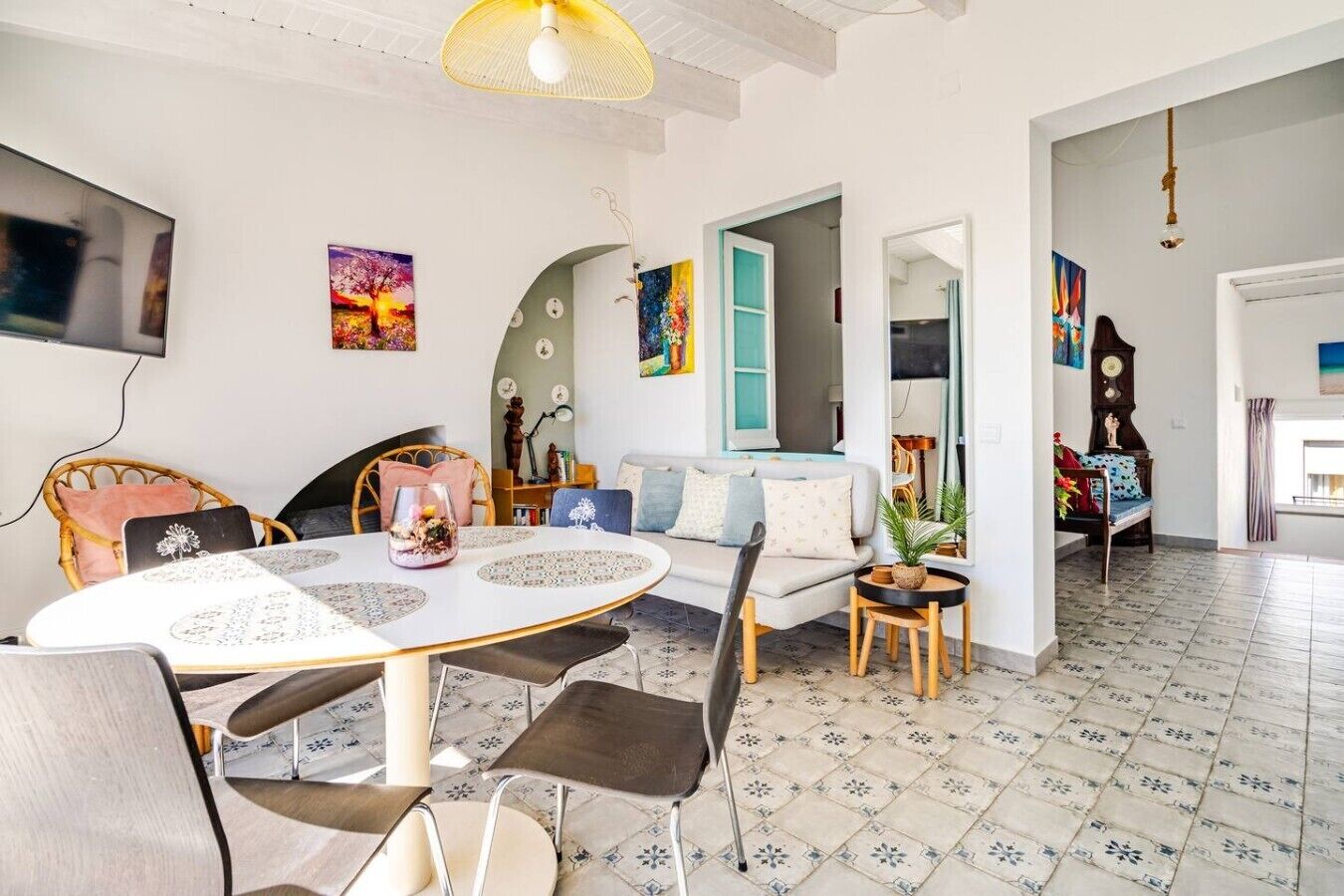 A modern living room with a round dining table, a sofa, and a coffee table. There is a TV mounted on the wall, and the room has a tiled floor.