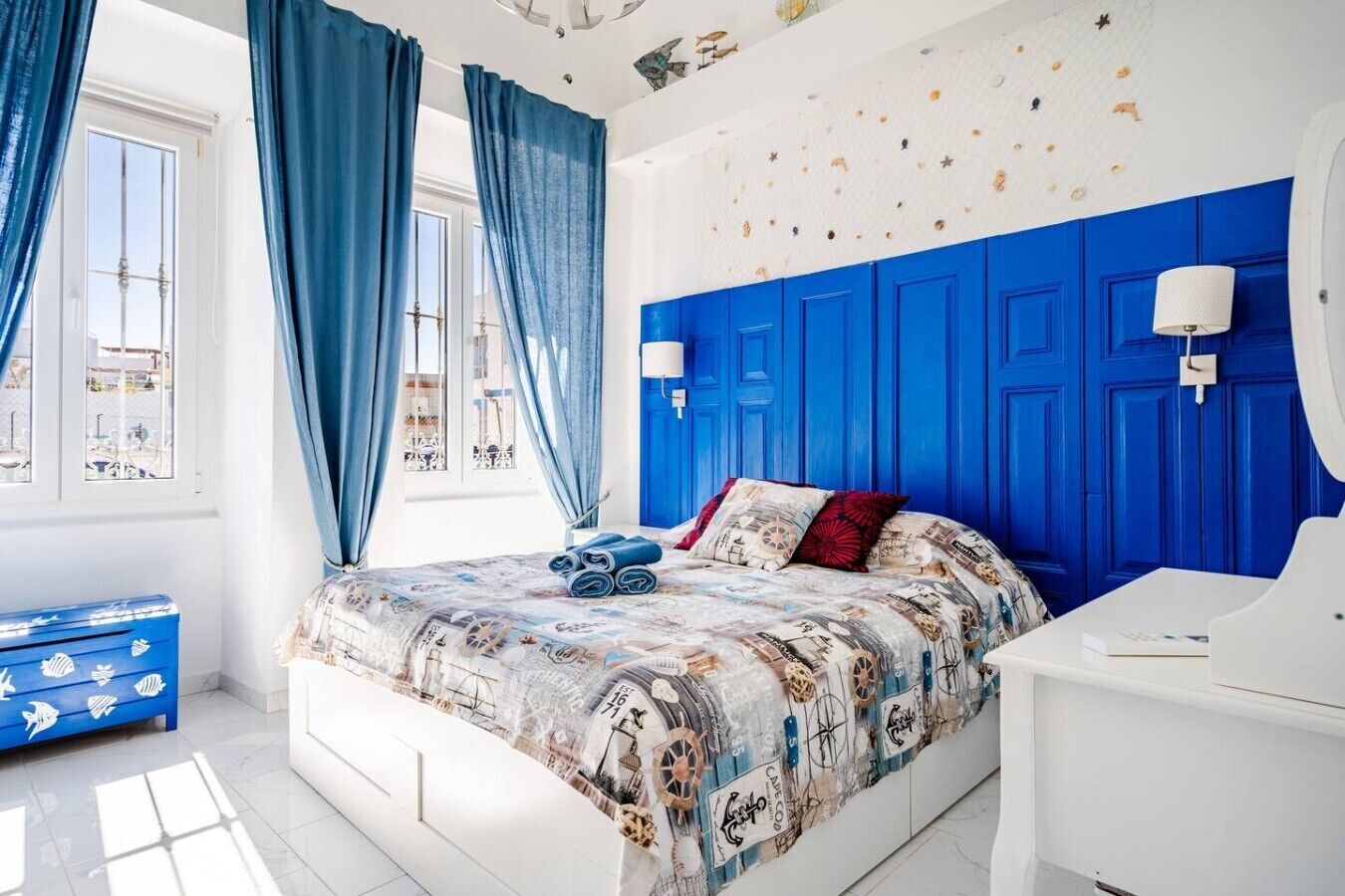 Ein Schlafzimmer mit blauem Kopfteil, einem Bett mit maritimer Bettwäsche, einer weißen Kommode und einem Fenster mit blauen Vorhängen.