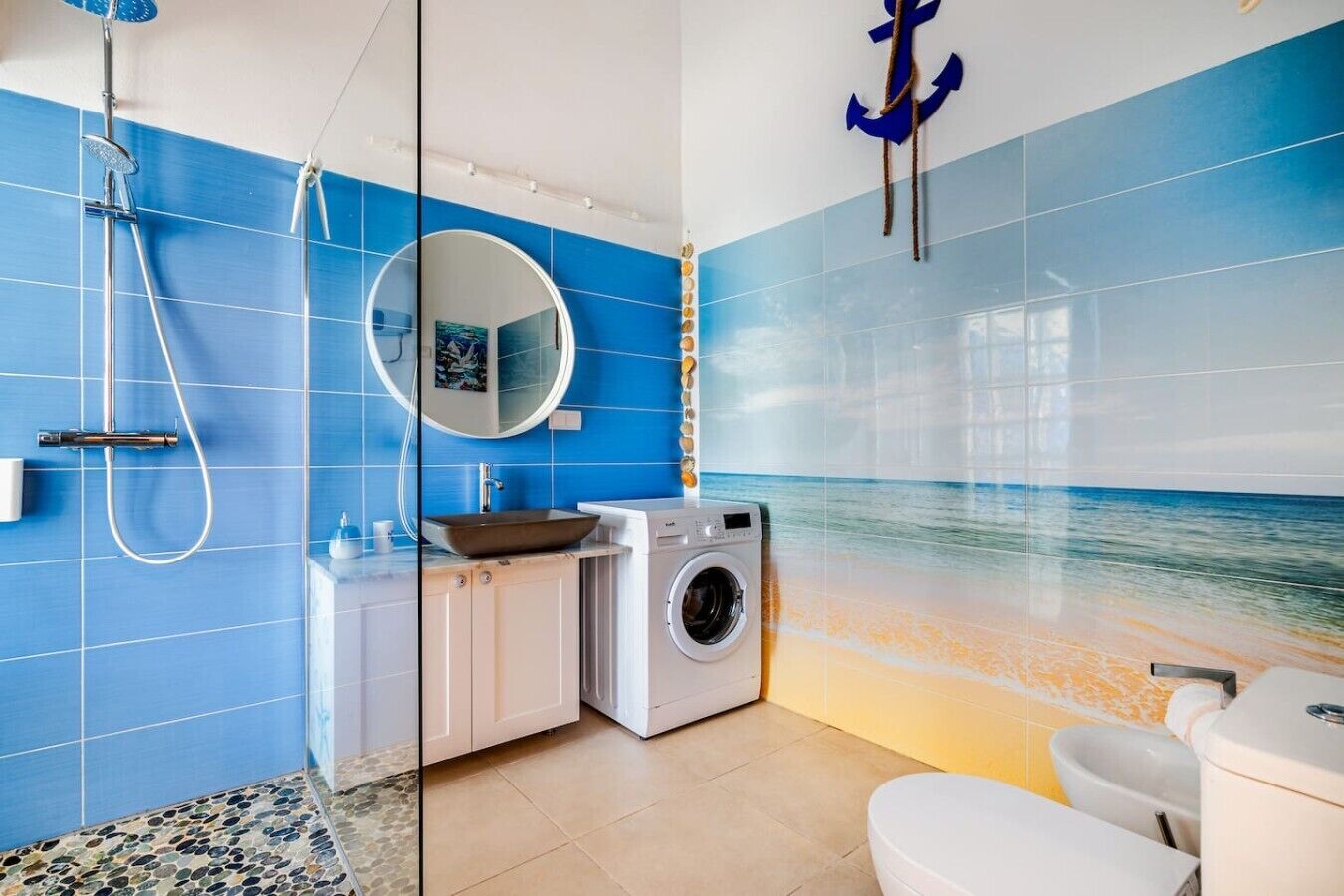 Een badkamer met een blauwe betegelde muur, een ronde spiegel, een wastafel, een wasmachine en een toilet.