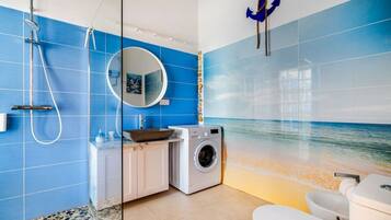 Een badkamer met een blauwe betegelde muur, een ronde spiegel, een wastafel, een wasmachine en een toilet.