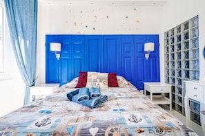 Quarto com cabeceira azul, cama com colcha de tema náutico, duas luminárias de cabeceira e uma mesa de cabeceira branca.