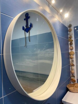 A bathroom with a large oval mirror reflecting a beach scene, blue tiles, and a wall-mounted shell display. - casa BOA VIDA, 150 square meters for YOUR comfort (Olhão)