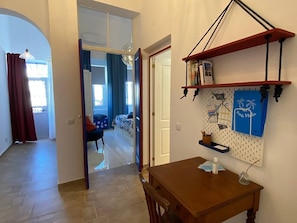 Quarto com mesa de jantar de madeira, painel mural com vários objetos e vista para outro cômodo com cama e cortinas.