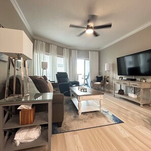 A modern living room with a ceiling fan, a flat-screen TV, a sofa, a coffee table, and a television stand. - Modern 1BR Beachfront Condo & Beautiful Courtyard Pool - D43 Orange Beach AL (Orange Beach)