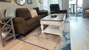 A living room with a brown sofa, a white coffee table, a round mirror, and a view of the outside through large windows.