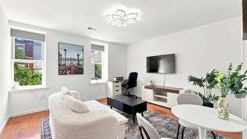 A modern living room with a sofa, a dining table, a TV, and a view of the outdoors through the windows.