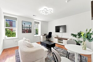 Ein modernes Wohnzimmer mit einer Couch, einem Esstisch, einem Fernseher und Blick ins Freie durch die Fenster.