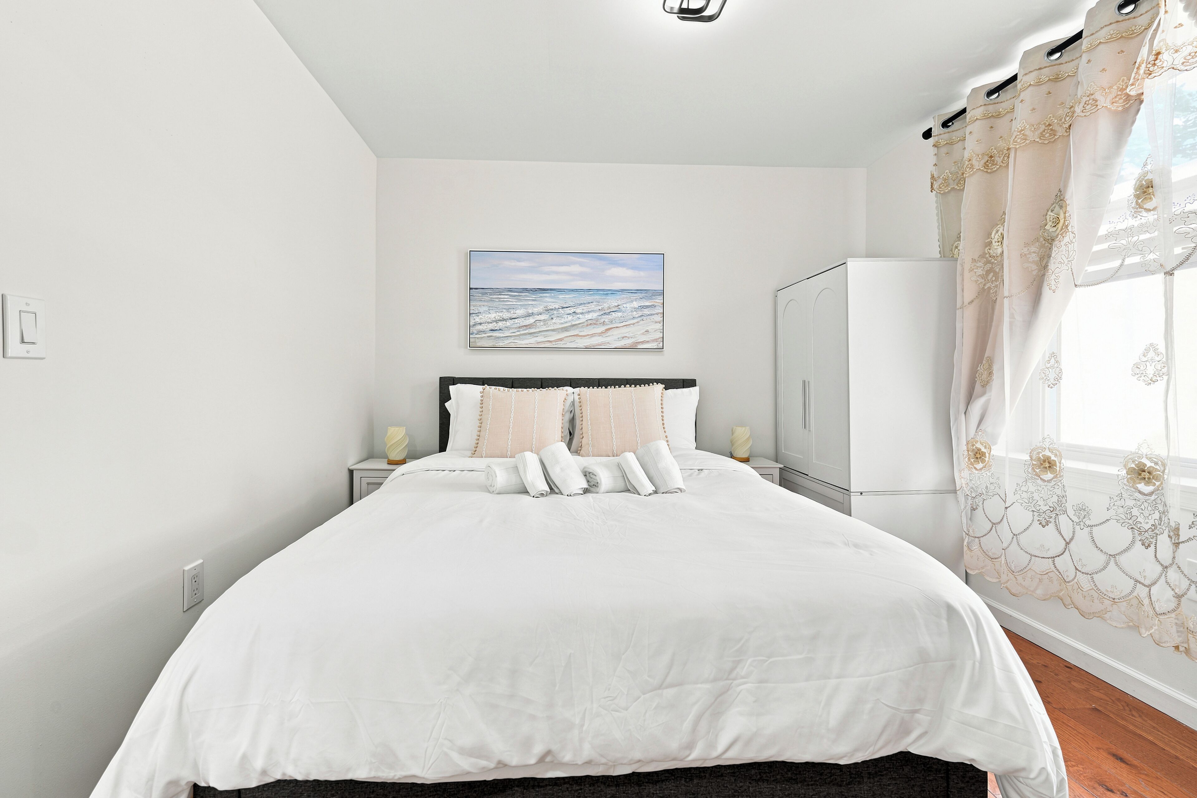 A bedroom with a bed, a nightstand, a wall-mounted picture, and a window with curtains.