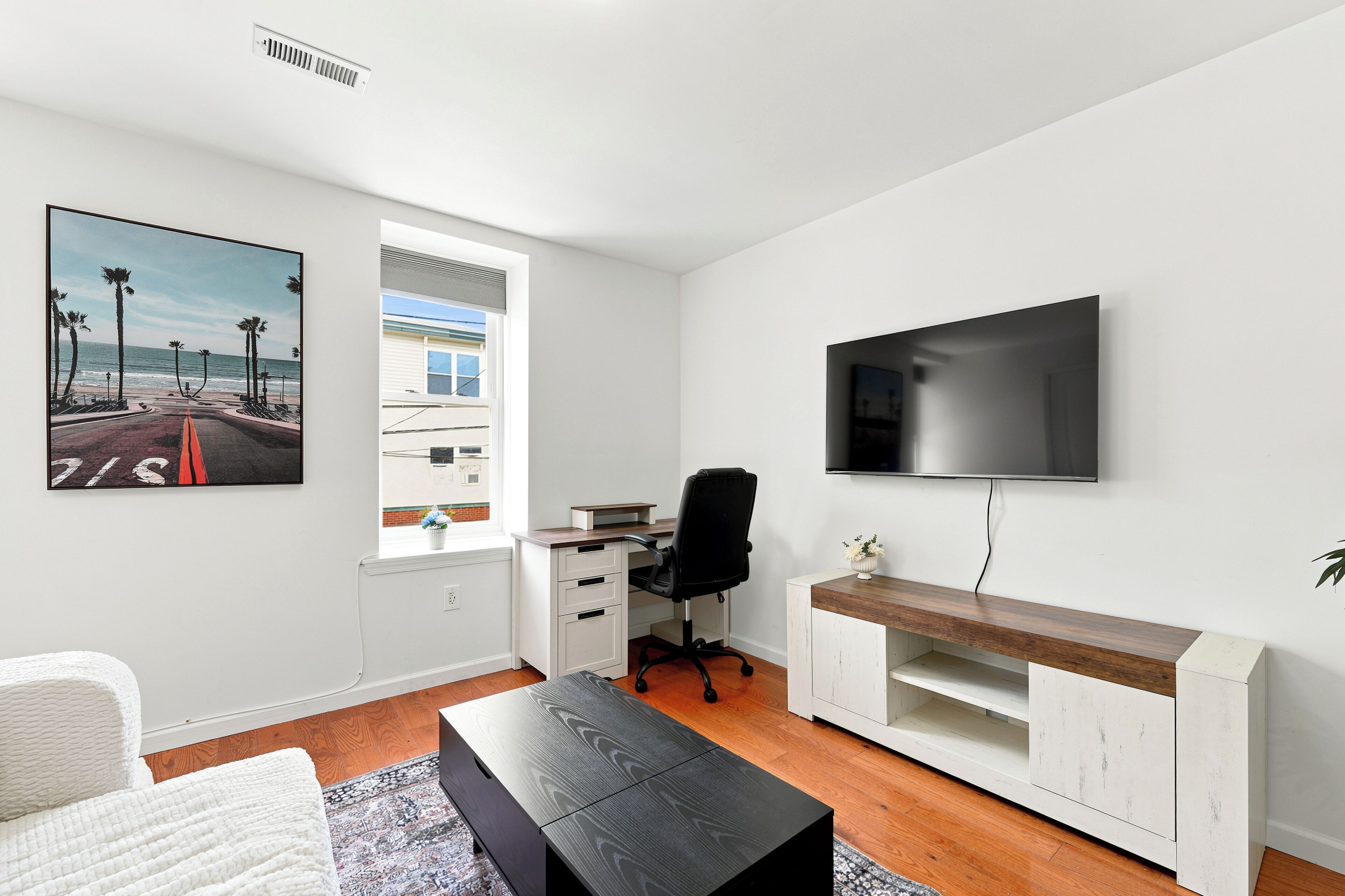 A modern living room with a flat-screen TV mounted on the wall, a desk with a chair, a sofa, and a large window with a view of palm trees and a beach.