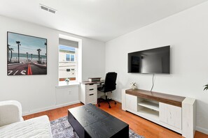 A modern living room with a flat-screen TV mounted on the wall, a desk with a chair, a sofa, and a large window with a view of palm trees and a beach. - Private 1-bed apartment w/ full kitchen & laundry in University City (Philadelphia)