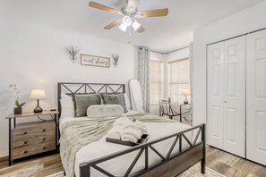 Quarto com cama, mesas de cabeceira, ventoinha no teto, placa na parede e janela com persianas.
