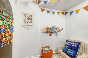 Quarto com parede de lousa, placa 'COOL KIDS CLUB', decoração com um carrinho de brinquedo colorido e cadeira de vime com almofada.