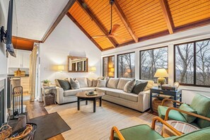 A spacious living room with a high ceiling, a fireplace, and large windows offering a view of trees.