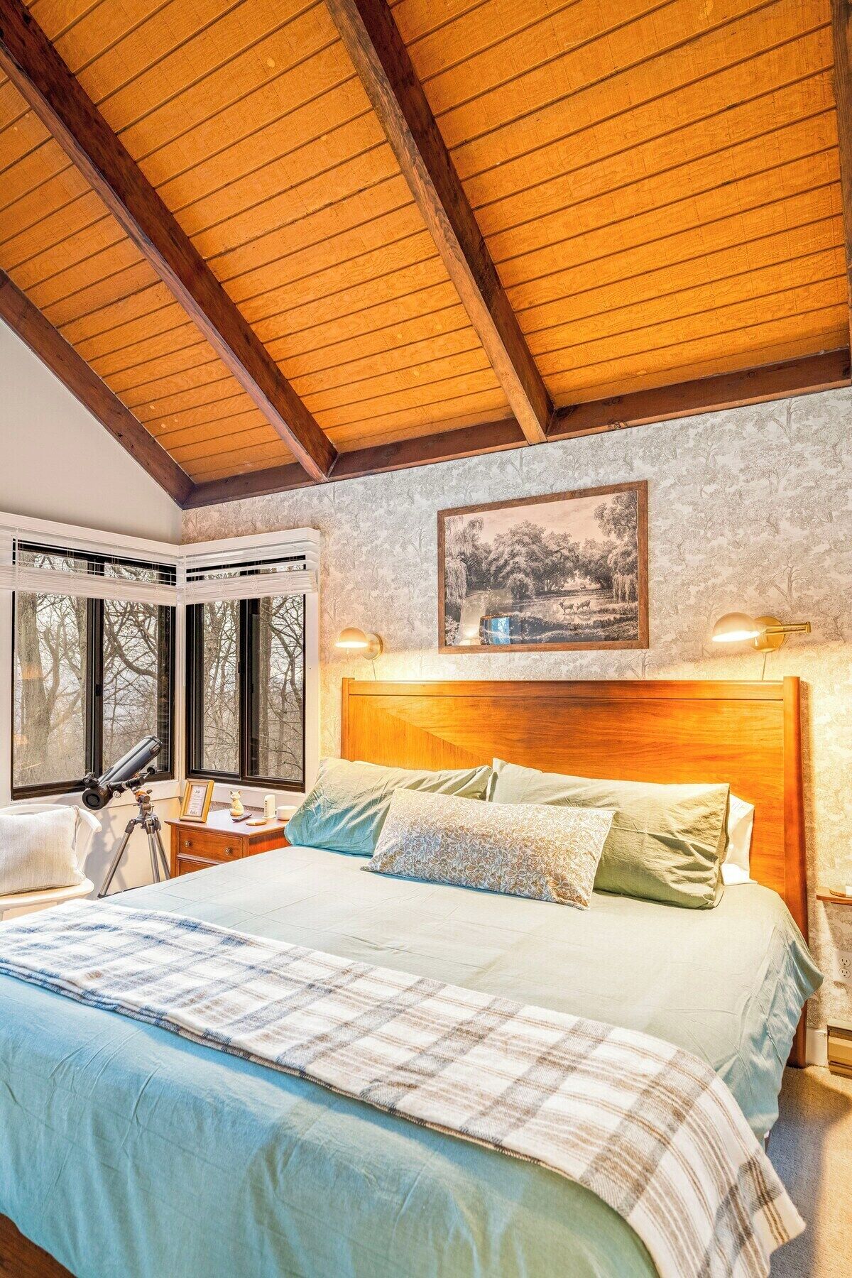 A bedroom with a large bed, wooden ceiling beams, a nightstand, a telescope, and a framed picture on the wall.