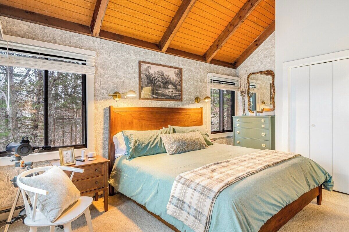 A bedroom with a wooden headboard, a bed with a plaid blanket, a nightstand with a lamp, a dresser, and a window with a view of trees.